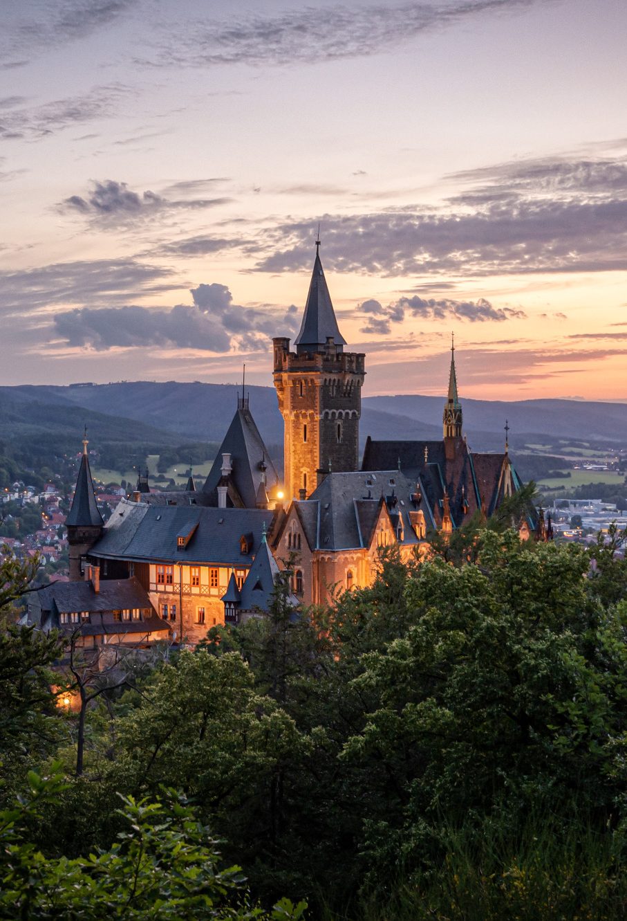 Blick auf eine beleuchtete Burg bei Sonnenuntergang, umgeben von Hügeln.